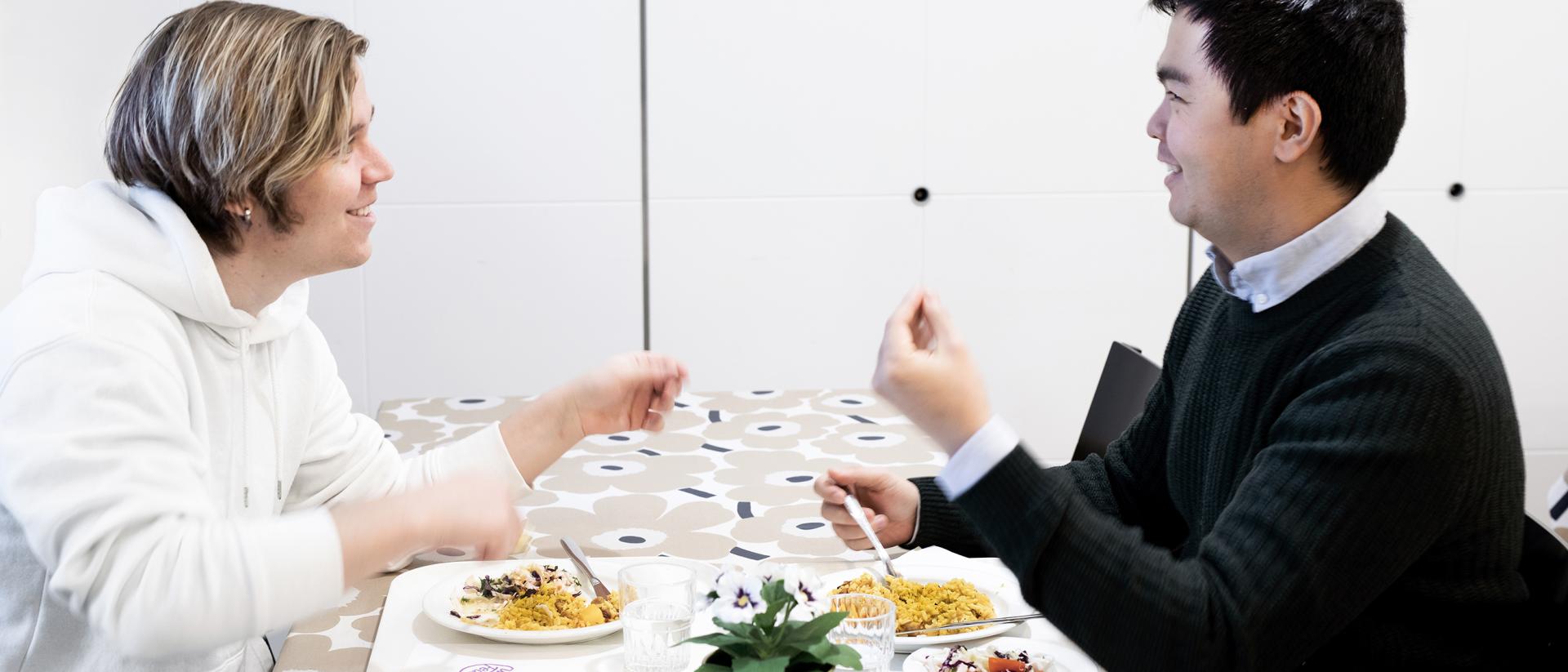 Kaksi henkilöä istuu syömässä ruokapöydän ääressä ruoka-annokset edessään