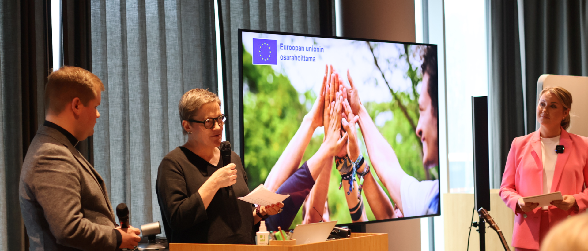 Kolme henkilöä luentosalin etuosassa, taustalla valkokankaalla iso valokuva yhteenliitetyistä käsistä