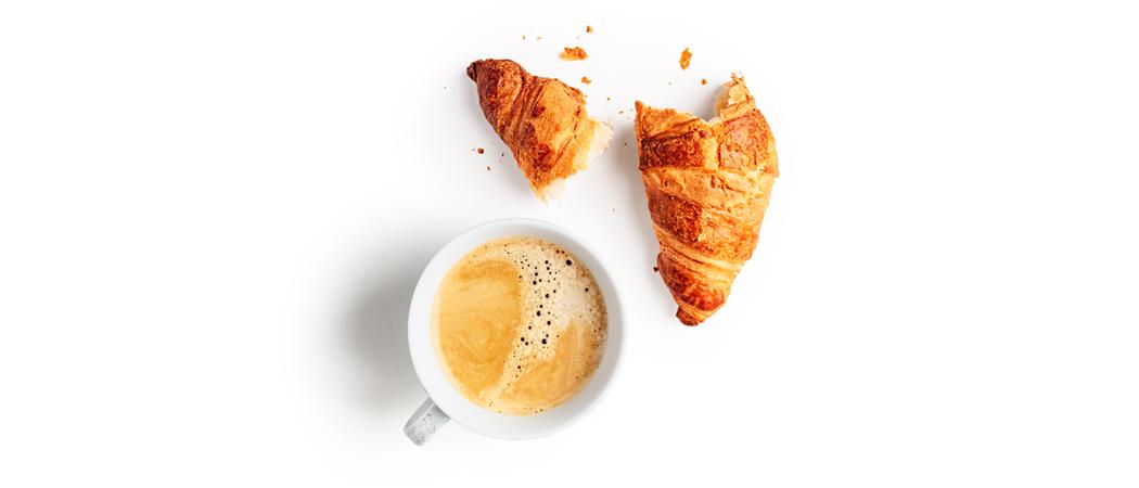 Valkoisella pöydällä halkaistu croissant ja muruja, sekä kuppi kahvia.