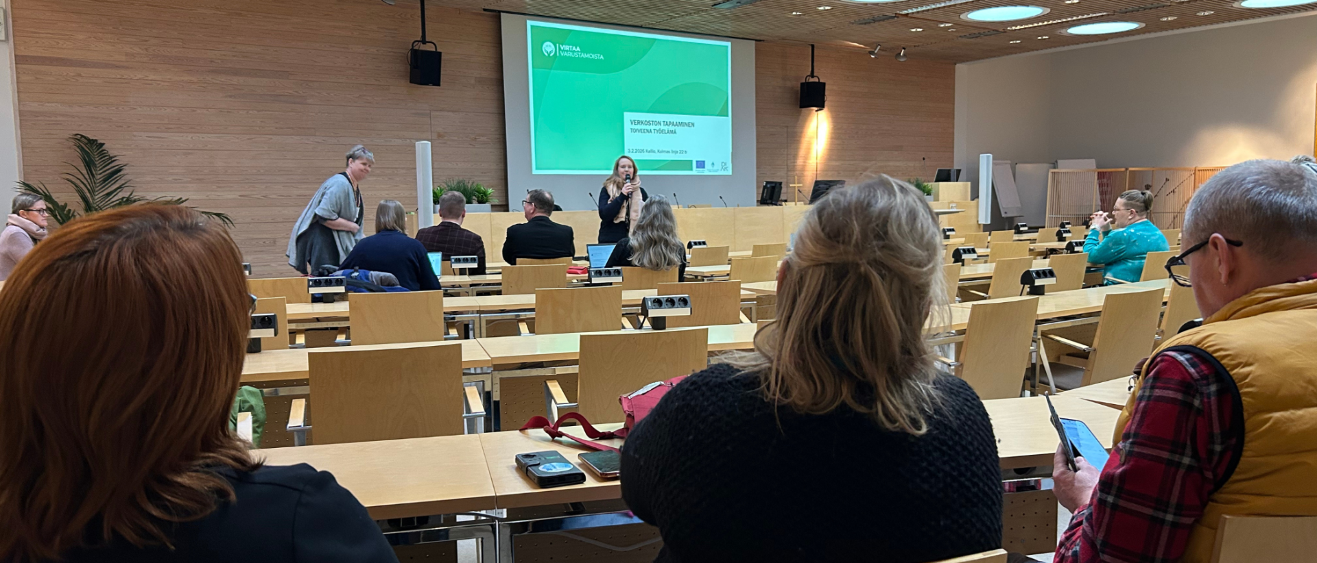 Luentosalissa istuu ihmisiä, edessä kuva isolla näytöllä.