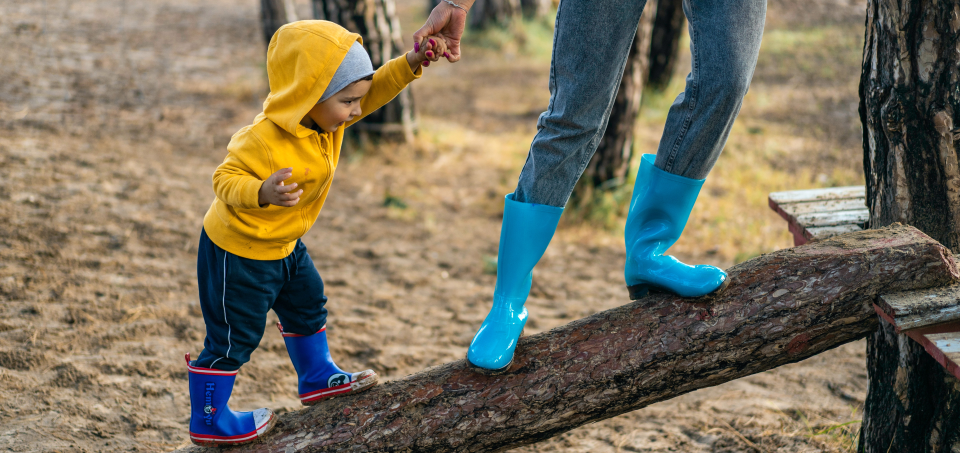 Lapsi kävelee värikkäissä sadevaatteissa kumisaappaat jalassa puunrungon päällä ja aikuinen pitää häntä kädestä kiinni.