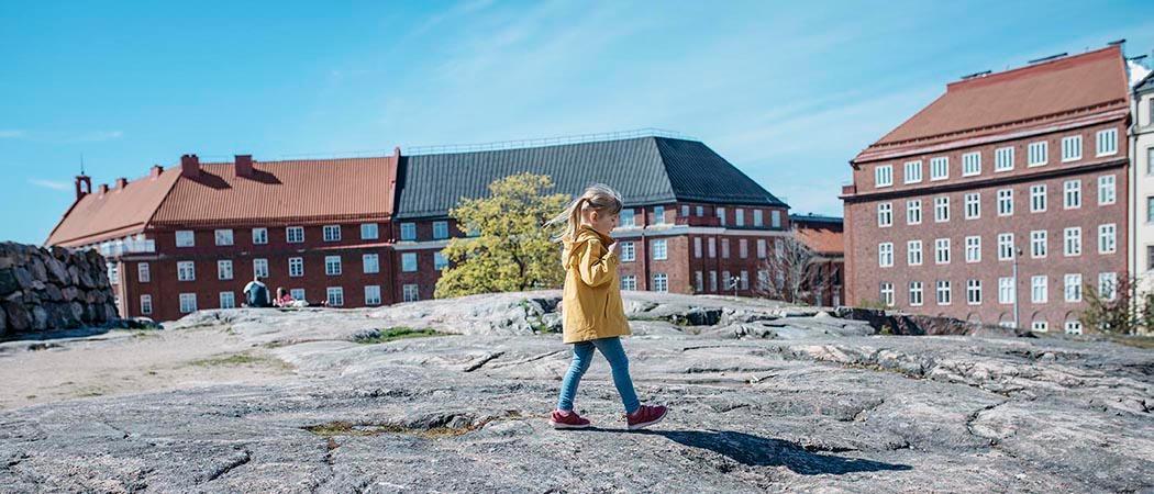 Keltatakkinen lapsi kävelee kalliolla, takana näkyy Töölön taloja. A child with a yellow coat walking on the rock, Töölö neighborhood's buildings on the background.