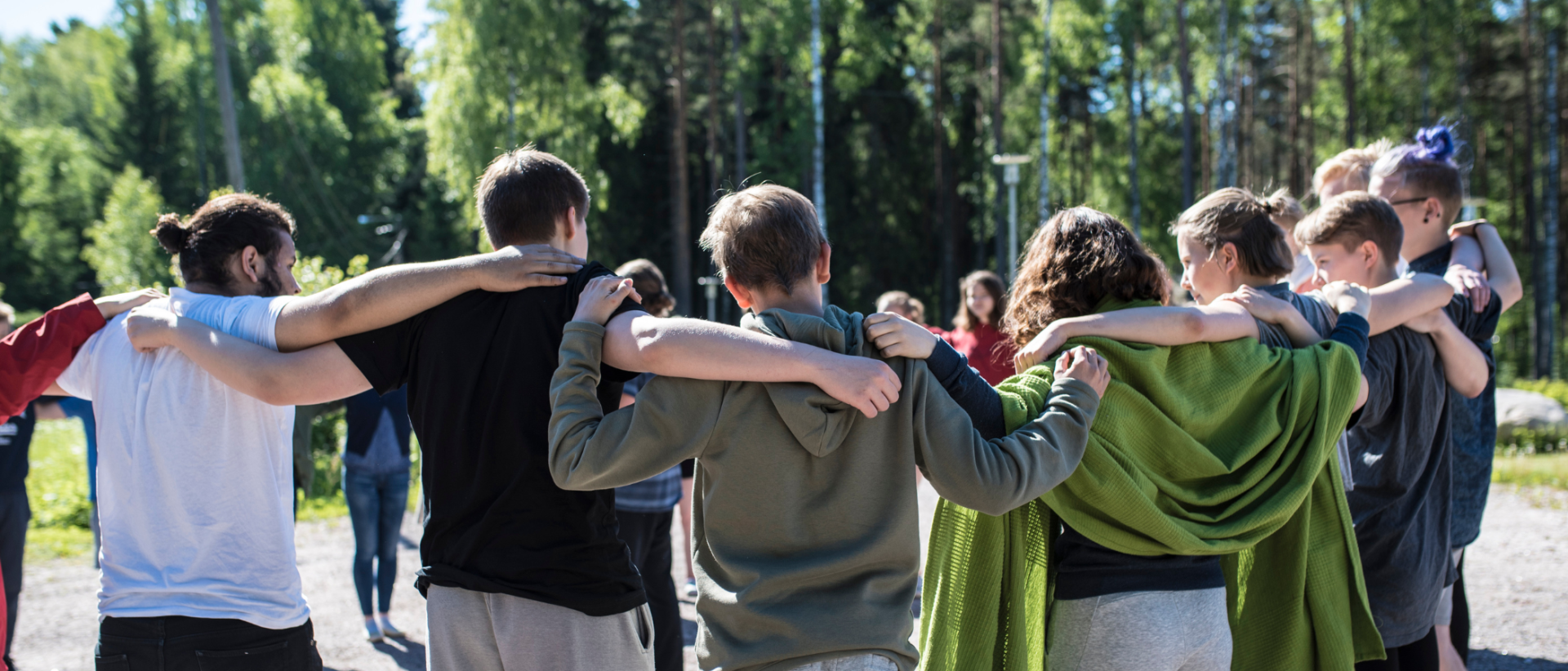 Kuvassa nuoria ulkona kesällä ringissä kädet toistensa harteilla, kuvattuna selkäpuiolelta.