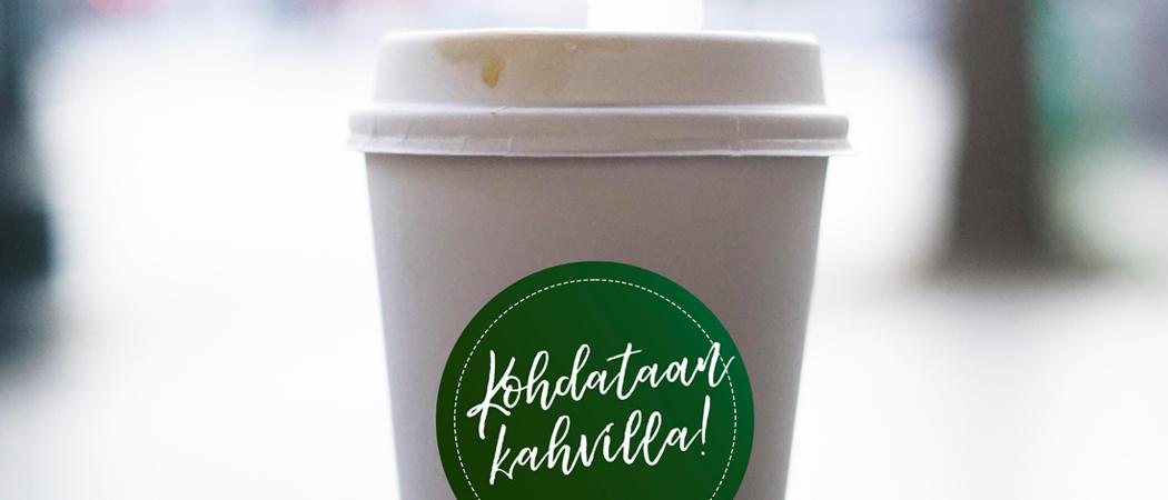 Valkoinen paperimuki, jossa vihreässä tarrassa teksti: kohdataan kahvilla!