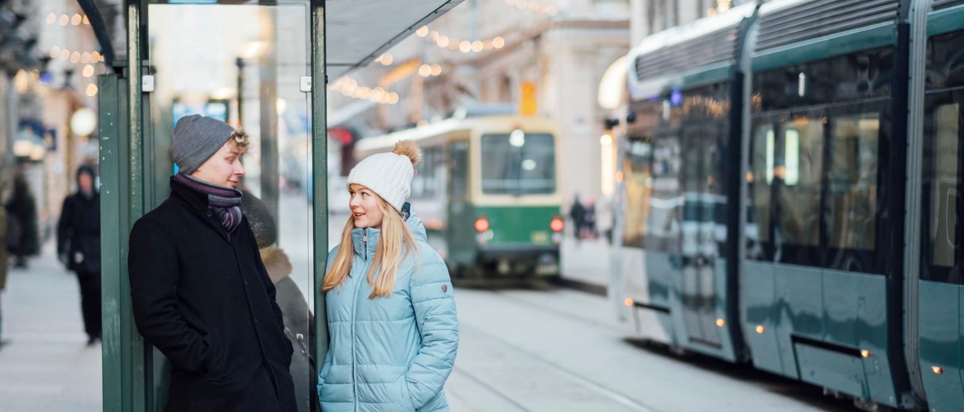 Kaksi nuorta seisoo raitiovaunupys&auml;kill&auml; juttelemassa, taustalla katun&auml;kym&auml; ja kaksi ratikkaa