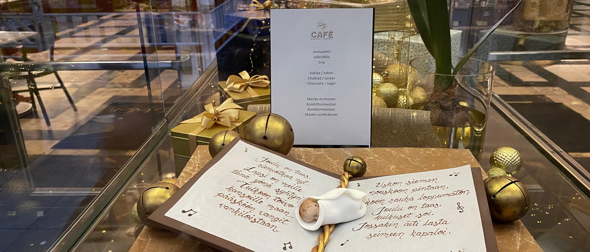 Suklaasta tehty seimiasetelma, jossa avonainen kirja ja Tulkoon joulu -laulun sanat sekä pieni vauvahahmo