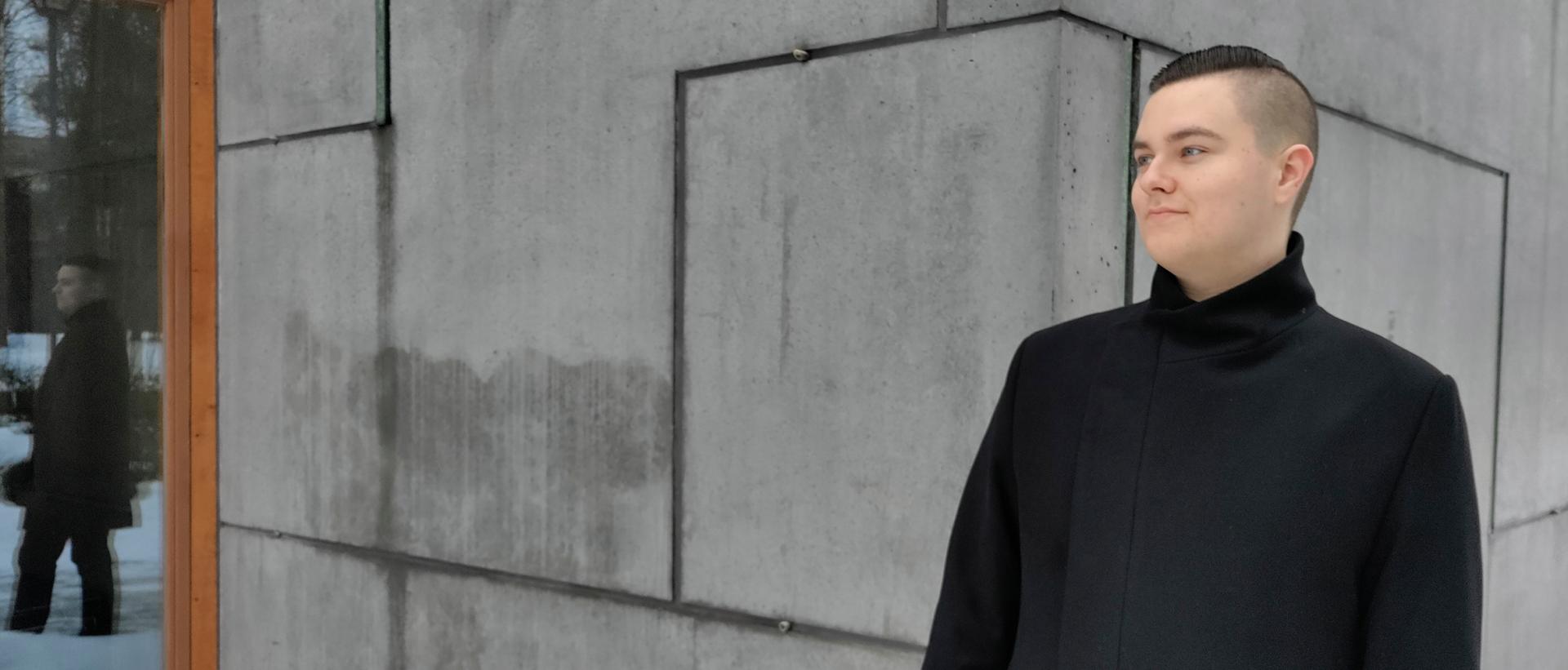 Teologian yo Antti Kosonen kuvattuna Laajasalon kirkon betoniseinää vasten tummassa takissa. Kuva: Remo Ronkainen