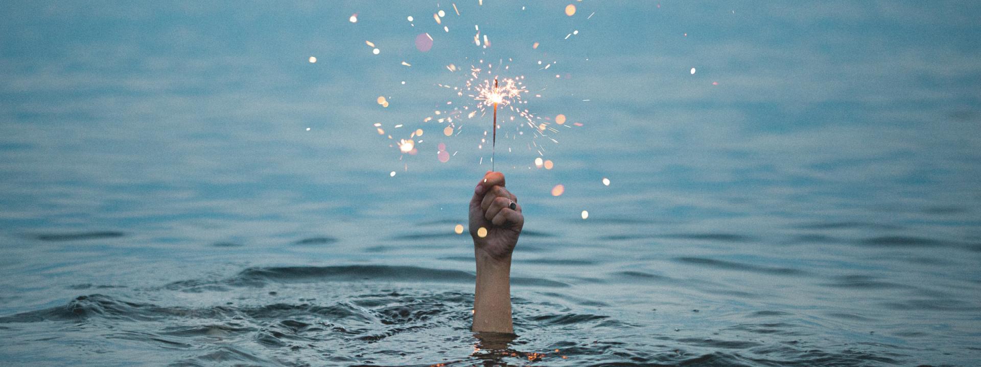 En hand sticker upp ur vattnet på en sjö, den håller ett brinnande tomtebloss.