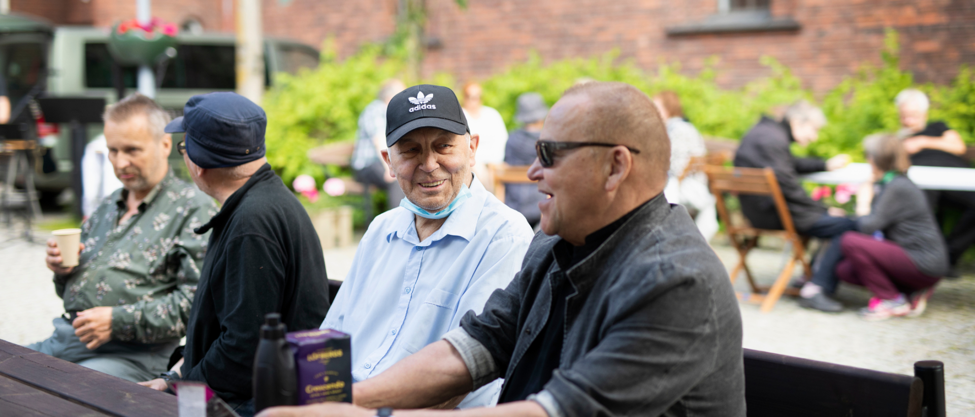 Ihmisiä istuu penkeillä kirkon pihalla juttelemassa, etualalla kaksi miestä, joista toisella vaalea paita ja lippis ja toisella tumma pusero ja aurinkolasit