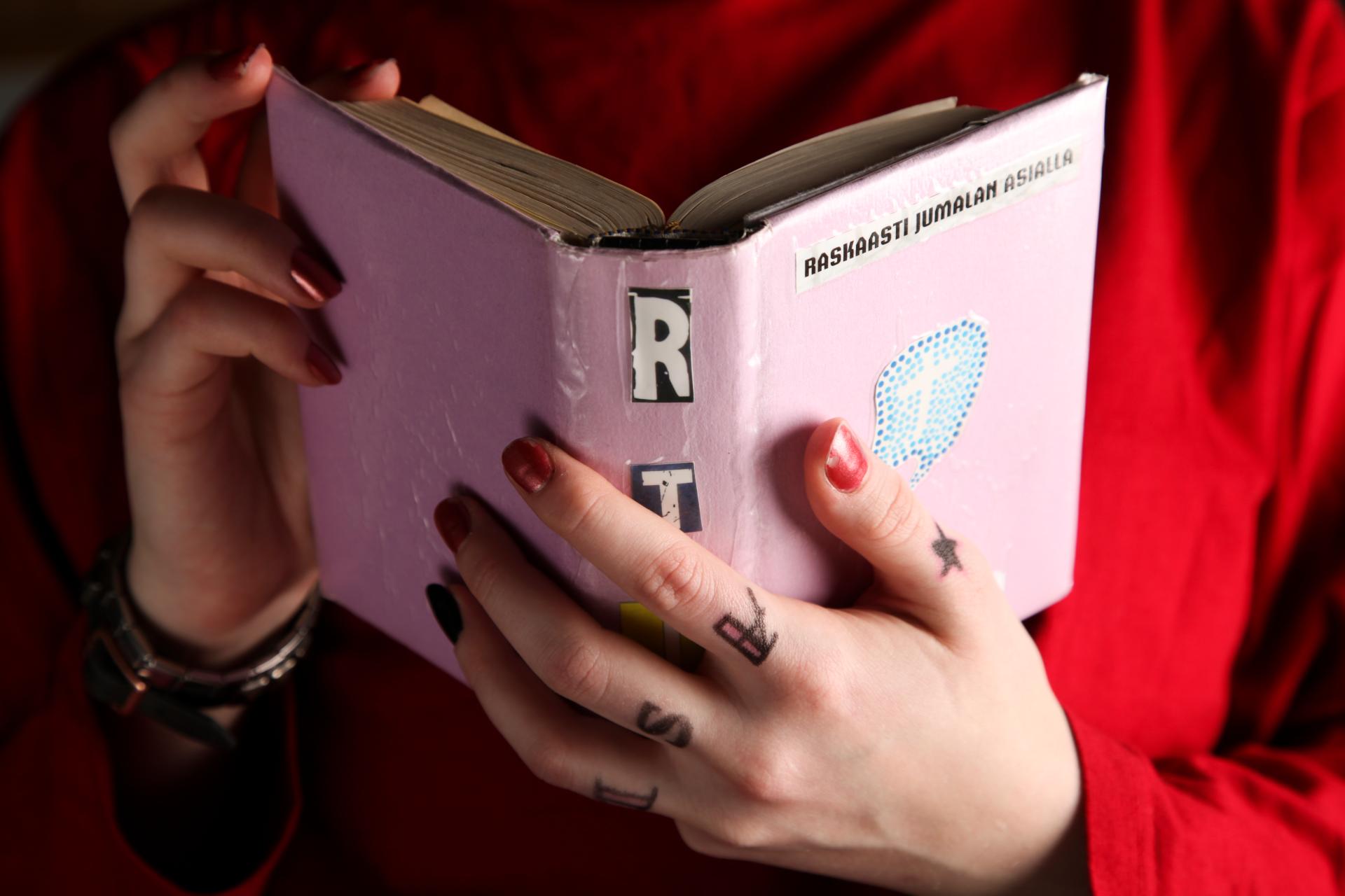 Puna-asuinen henkilö, jolla tatuoinnit sormissa ja kynsilakkaa, pitelee kädessään vaaleanpunakantista Raamattua