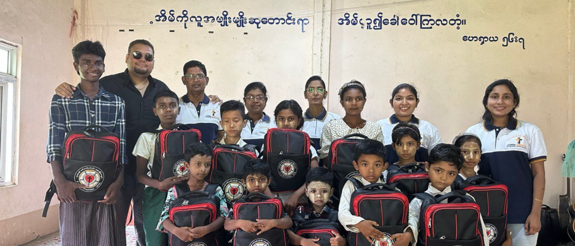 Myanmarilaisia lapsia seisoo riveissä koulureput sylissään, taustalla opettajia