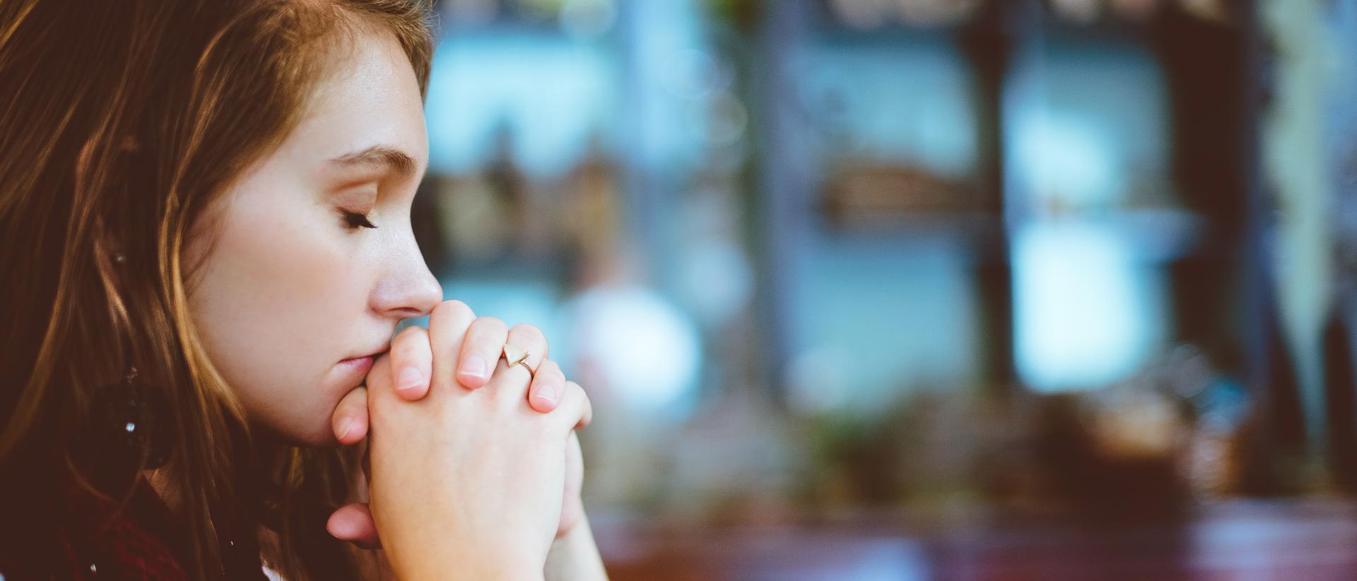 Nuori nainen istuu silmät kiinni ja kädet ristissä kasvojen edessä rukoilemassa