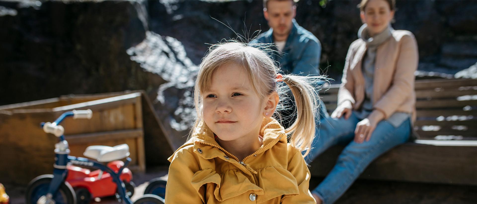 Etualalla saparopäinen pikkutyttö keltaisessa takissa. Takana vanhemmat istuvat hiekkalaatikon reunalla. Kuva: Elina Manninen
