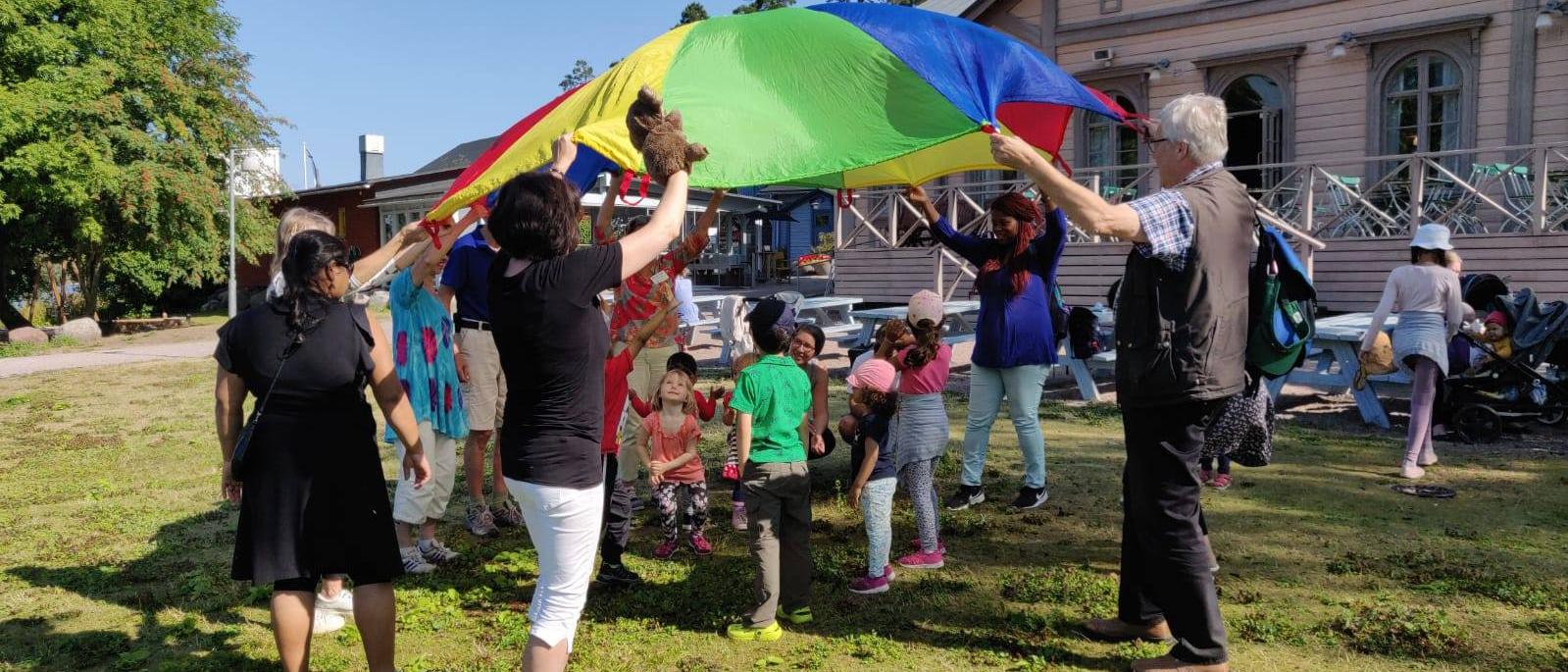 Kuva monikulttuuriselta retkeltä Mustasaaresta, jossa lapsia  ja aikuisia leikkimässä varjon alla.